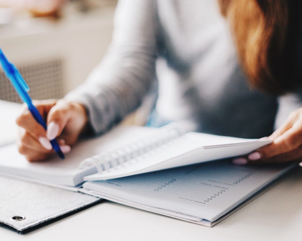 A woman with long hair sitting at a table writing with a blue pen in a spiral bound notebook.