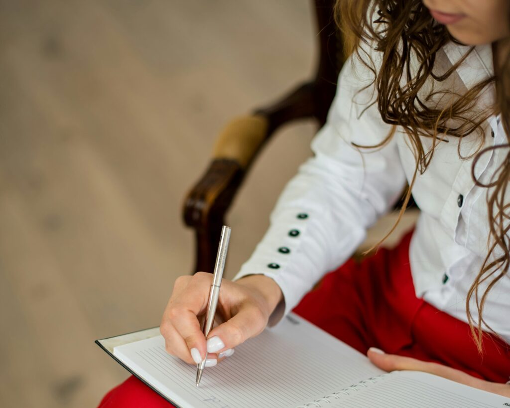 woman in white shirt and red pants sitting down and writing in a notebook