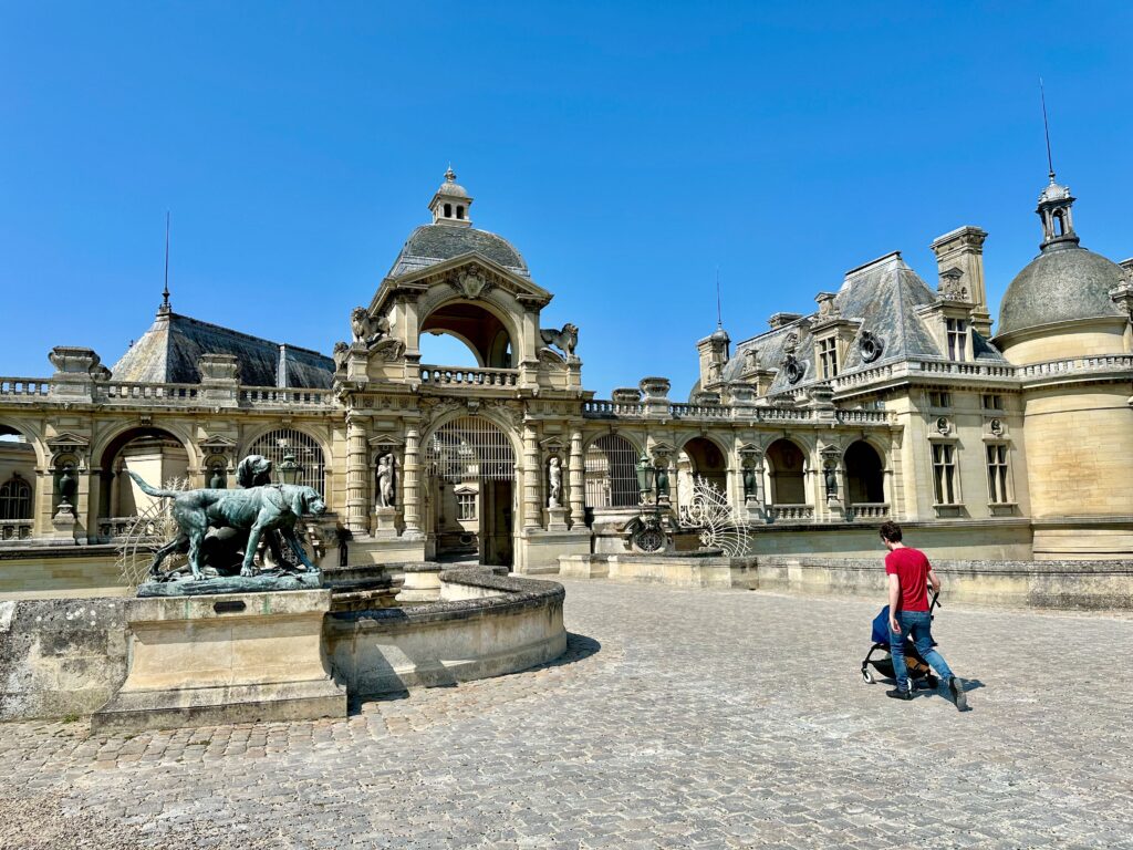 Man walking towards Entrance to Château de Chantilly and pushing a stroller over cobblestone pathway